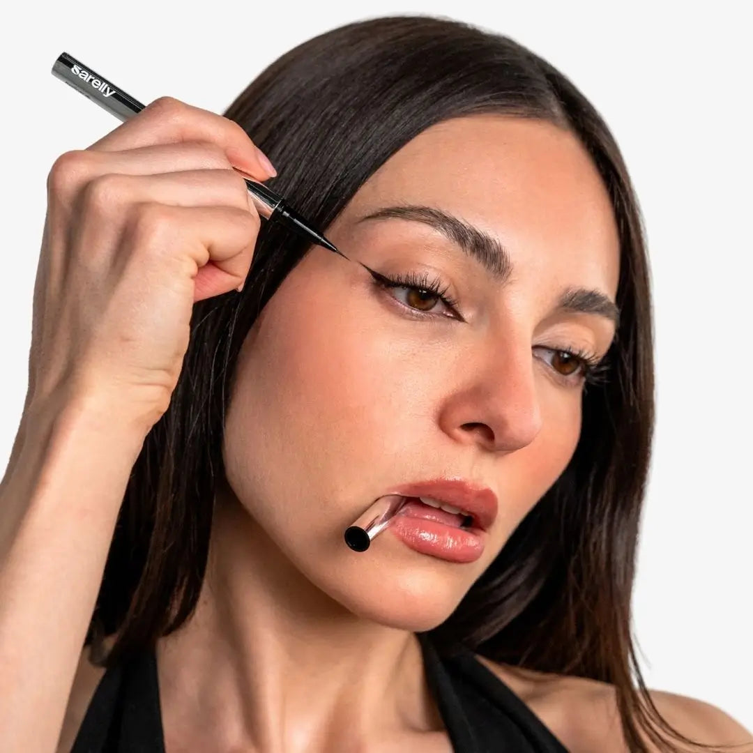 Close-up of a woman applying black eyeliner with a fine-tip pen while wearing a black halter top against a light gray background.