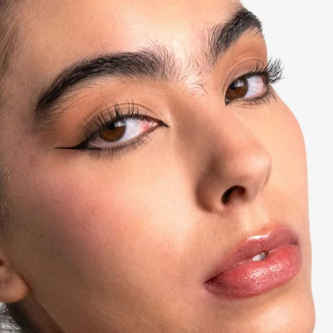 Close-up of a model's face showcasing winged black eyeliner, long eyelashes, and glossy pinkish-brown lips against a clean white background.