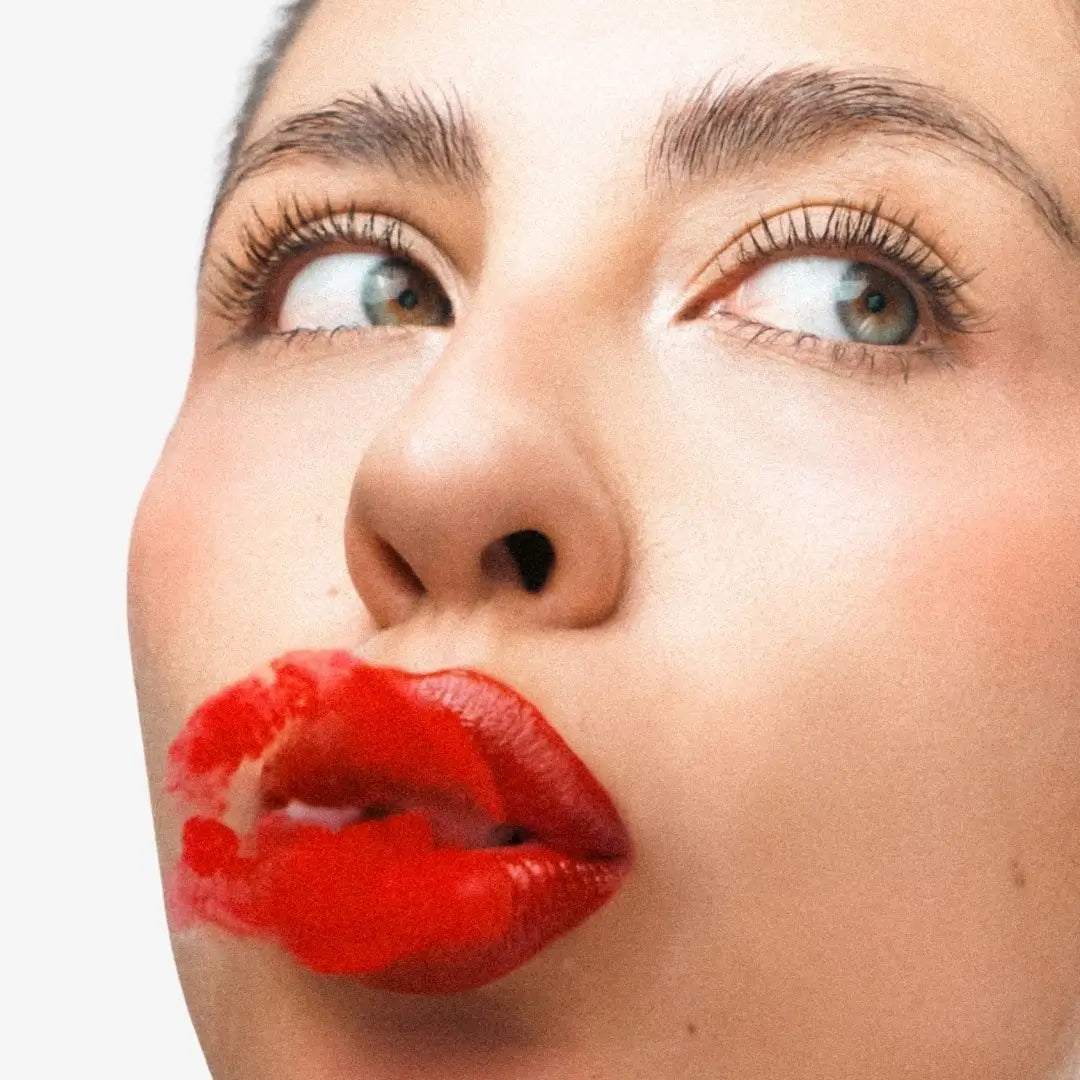 Close-up of a woman's face with bright red lipstick on her lips, looking upward against a plain light background. Shade Rubi