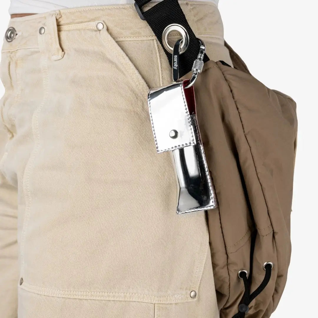 A close-up of a person wearing beige canvas pants and a brown backpack, with a shiny silver metallic pouch attached to a black strap on the pants. Shade Rubi