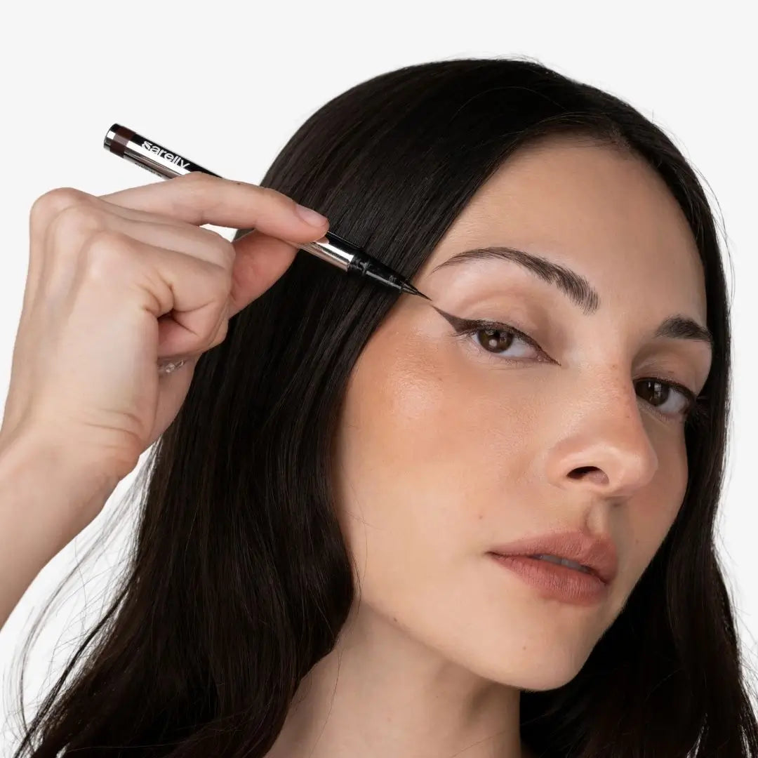 A close-up of a woman with long dark hair applying chocolate eyeliner with a fine-tip pen to her eyebrow, shown against a plain white background.