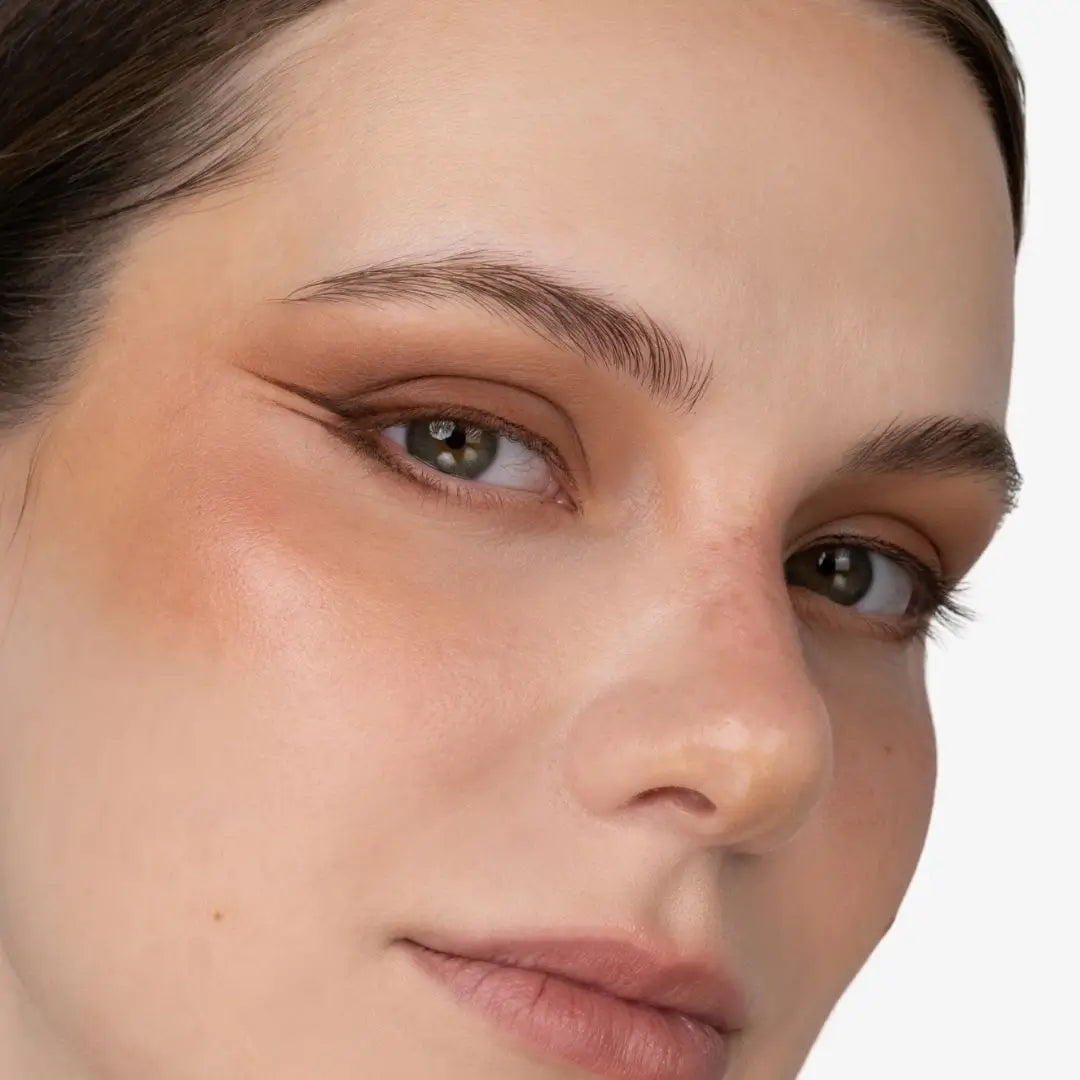 Close-up of a woman's face showing a warm, shimmery eyeshadow and blush makeup look on a clean white background. Shade Chocolate