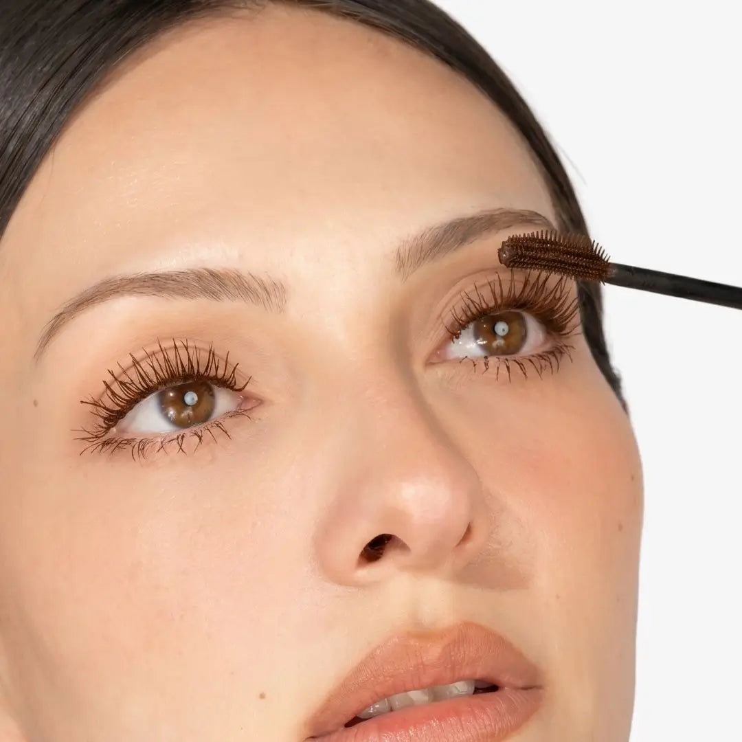 Close-up of a woman applying chocolate mascara to her eyelashes with a mascara wand against a clean white background.
