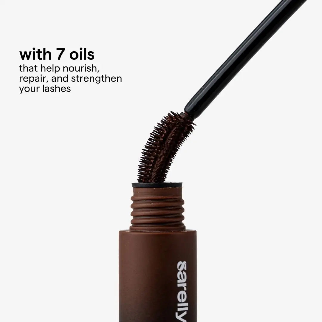 A close-up of a chocolate cosmetic tube with a applicator wand showing bristles, with text reading 'with 7 oils that help nourish, repair, and strengthen your lashes' on a light gray background.