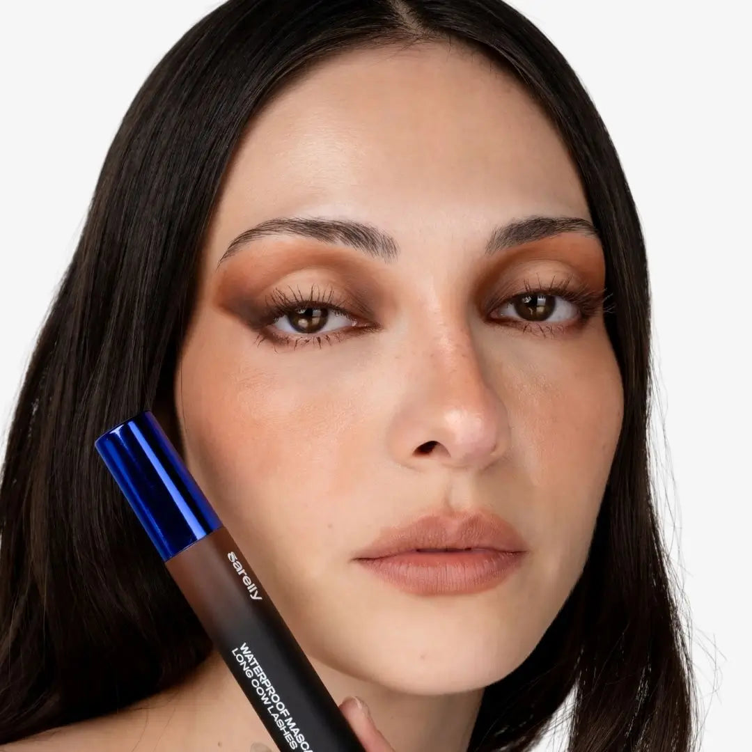 A close-up of a woman holding a cow lashes mascara chocolate with a metallic blue cap, showing the product against her face on a light gray background.