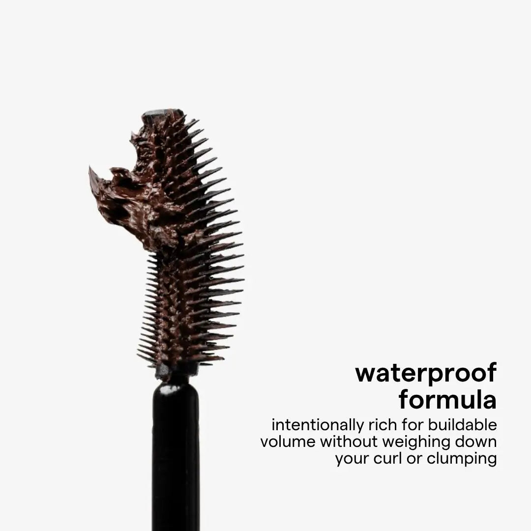 A close-up of a mascara wand showing a heavily coated, clumpy dark chocolate formula on the brush tip, with text on the right reading 'waterproof formula' and a smaller descriptive line, on a light gray background.