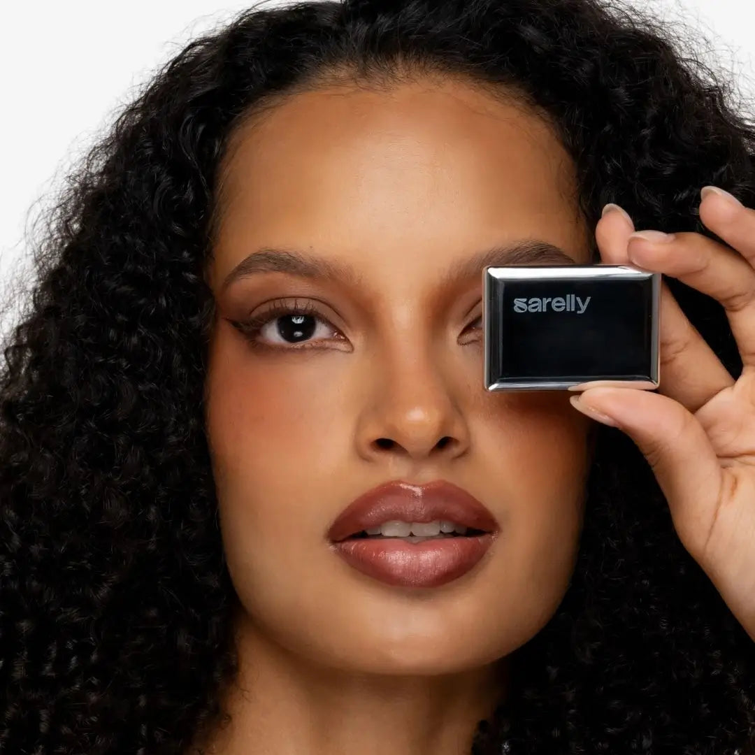A close-up of a woman holding a small rectangular black compact labeled 'sarelly' against her eye, with curly dark hair and a clean white background. Creamy Chocolate