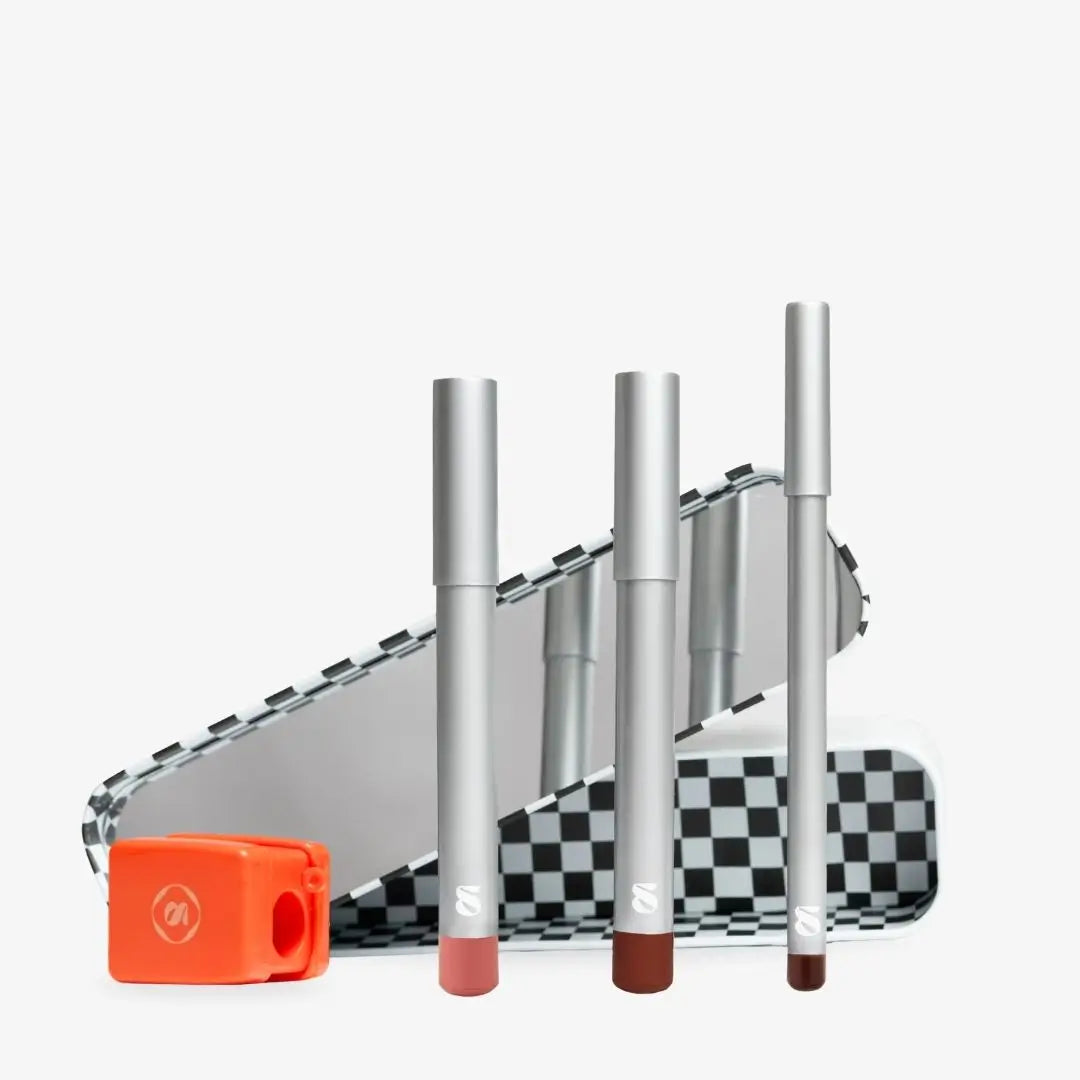 Three silver cosmetic tubes with pink and brown tips stand in front of a black-and-white checkered case, with an orange pencil sharpener on a light gray background. Shade chocolate
