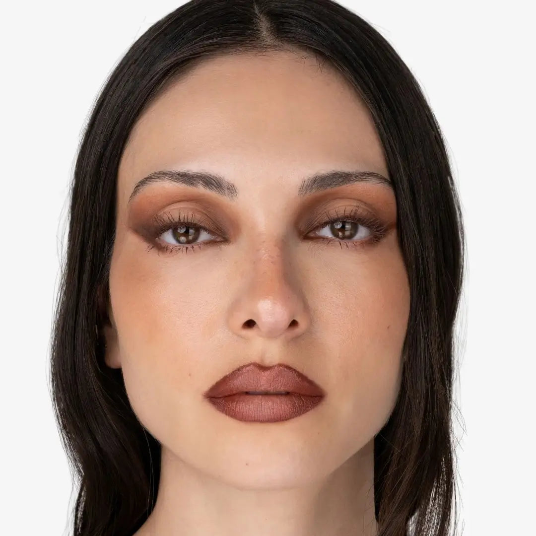 Close-up of a woman with makeup featuring warm brown eyeshadow and a muted brown lipstick, shown against a plain light gray background. Shade chocolate