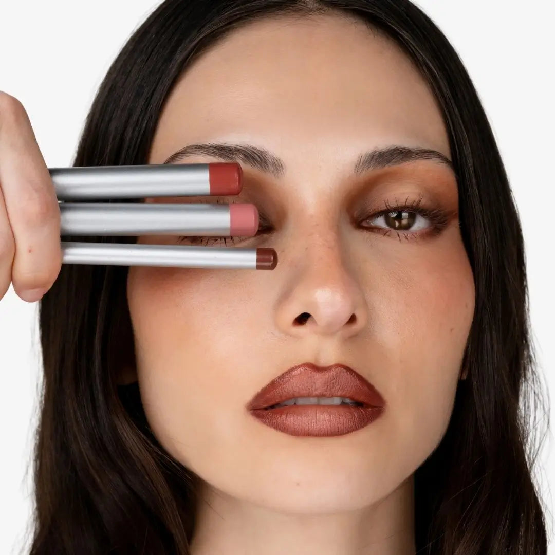 Close-up of a woman holding three makeup lip pencils against her eye, showing the tips of the pencils in different nude shades against a clean white background. Shade chocolate