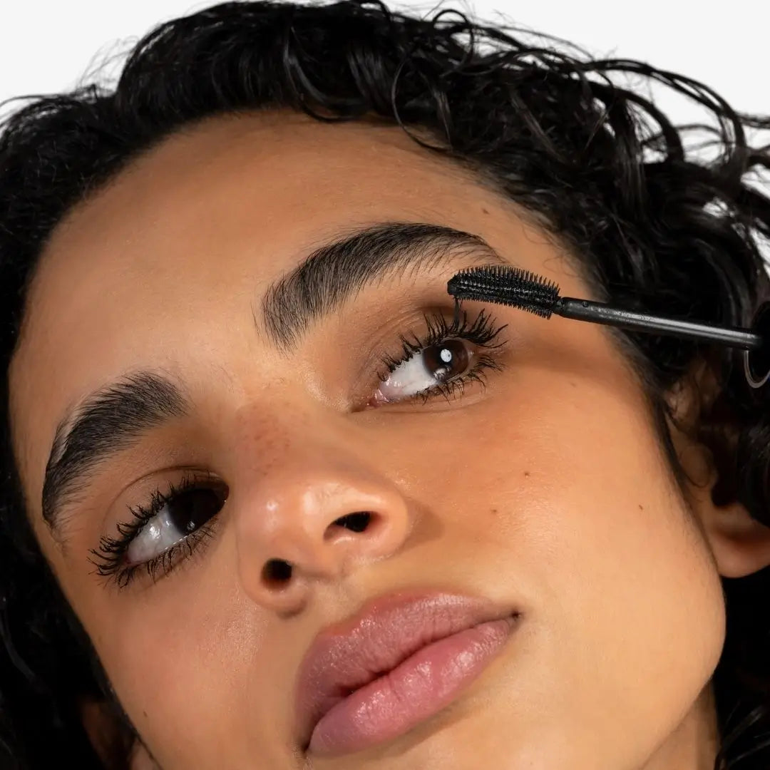 Close-up of a person applying mascara  cow lashes black to their eyebrows with the wand visible against a clean white background.