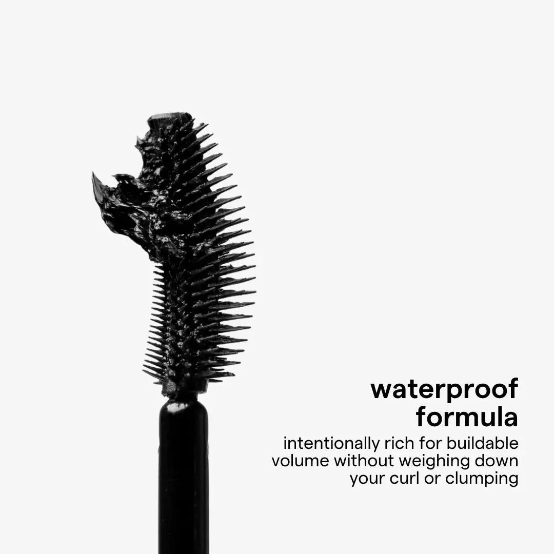 A close-up of a black mascara wand showing a thick, coated brush bristle on a light gray background, with Spanish text reading 'fórmula waterproof' and smaller descriptive copy.