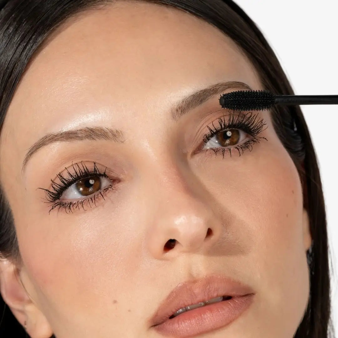 Close-up of a woman applying black mascara with a wand to her upper lashes against a clean white background.
