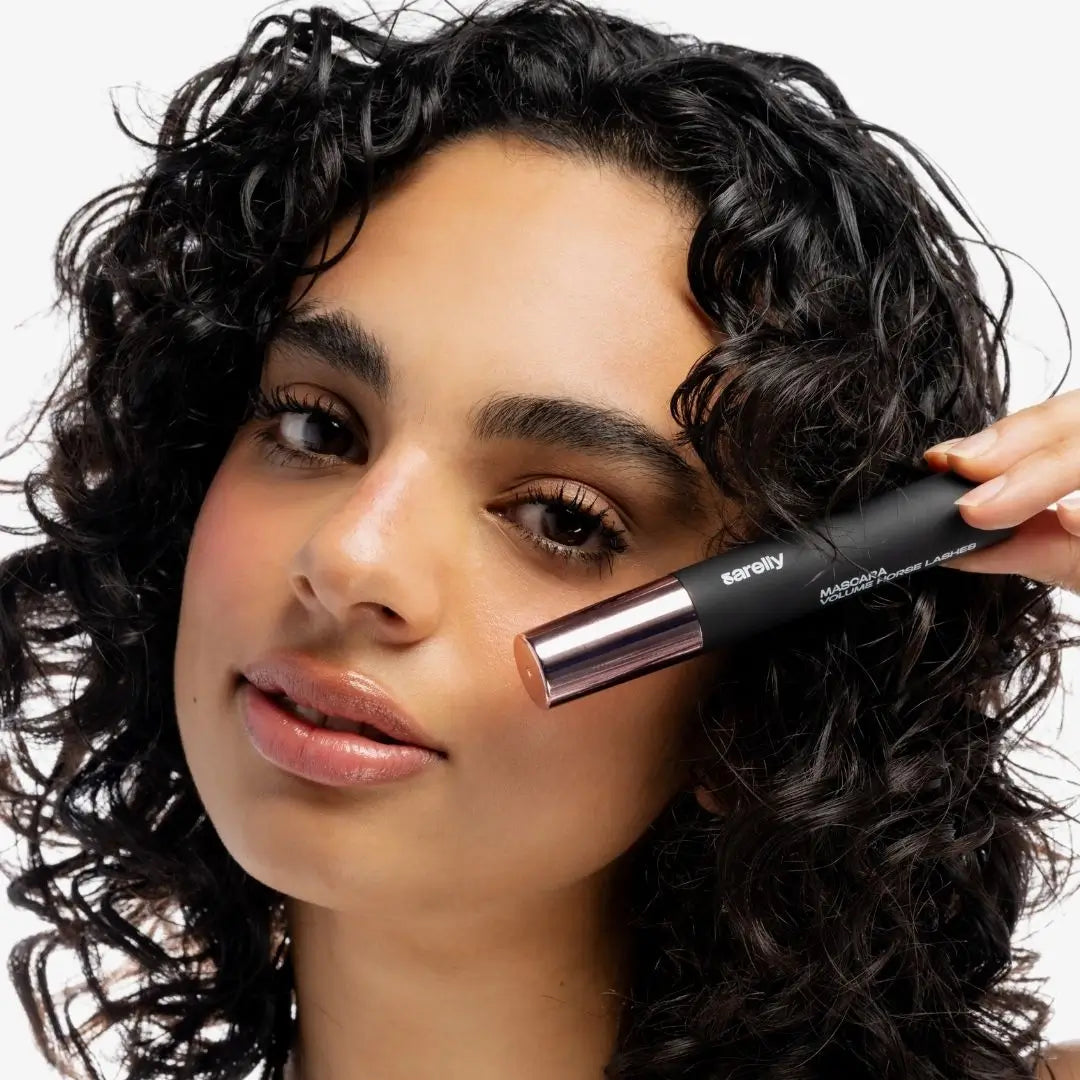 A close-up of a woman with curly dark hair showing the makeup product being applied, on a clean white background. Using horse lashes mascara black