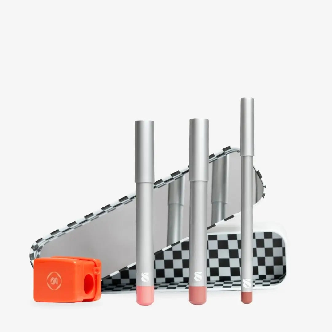 Four silver cosmetic tubes with pink and nude tips arranged in front of a black-and-white checkered case, with an orange compact case on a light gray background. Shade Creamy