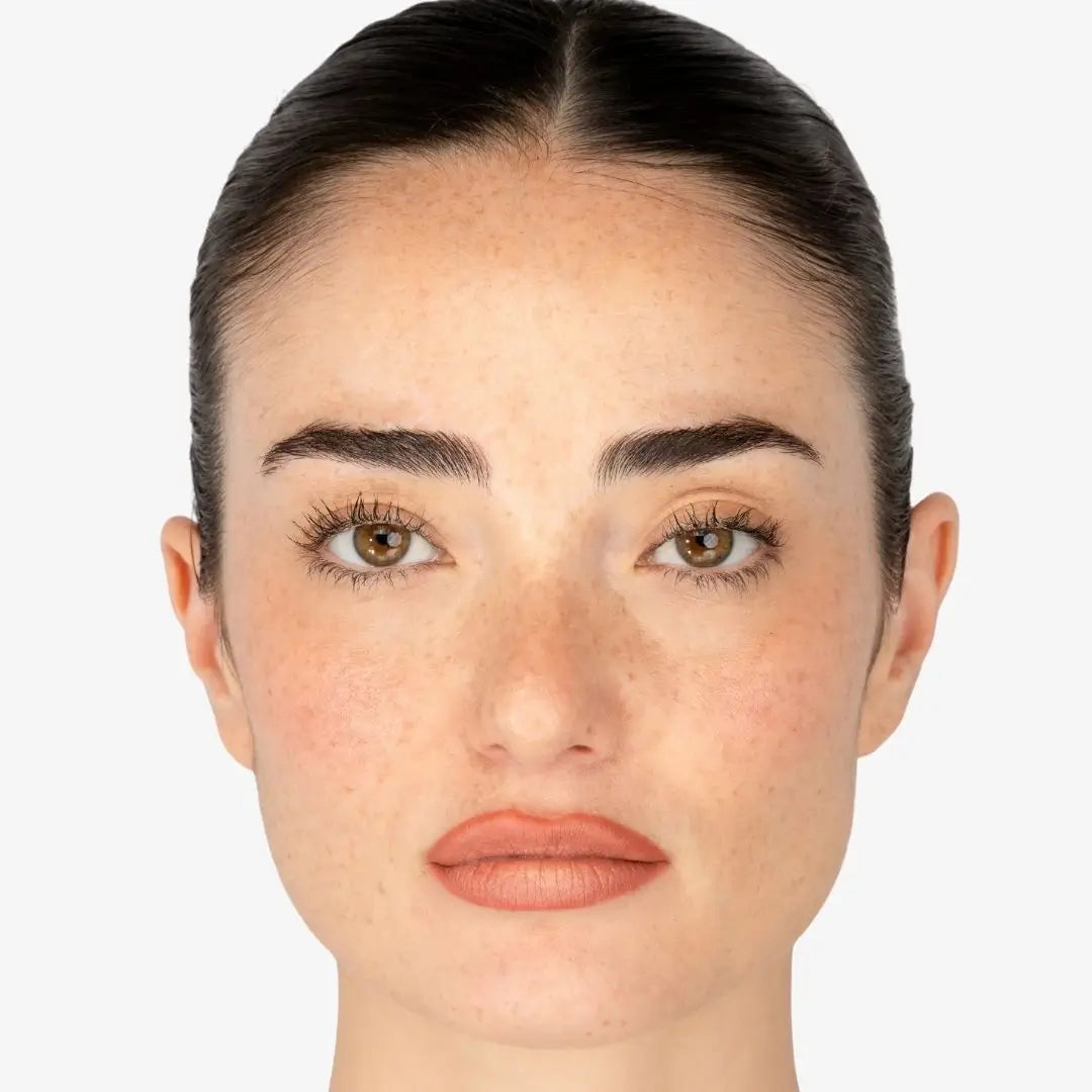 A close-up portrait of a woman's face with dark brown hair pulled back, brown eyes, freckles, and lip combo buchon Shade Creamy, shown against a light gray background.