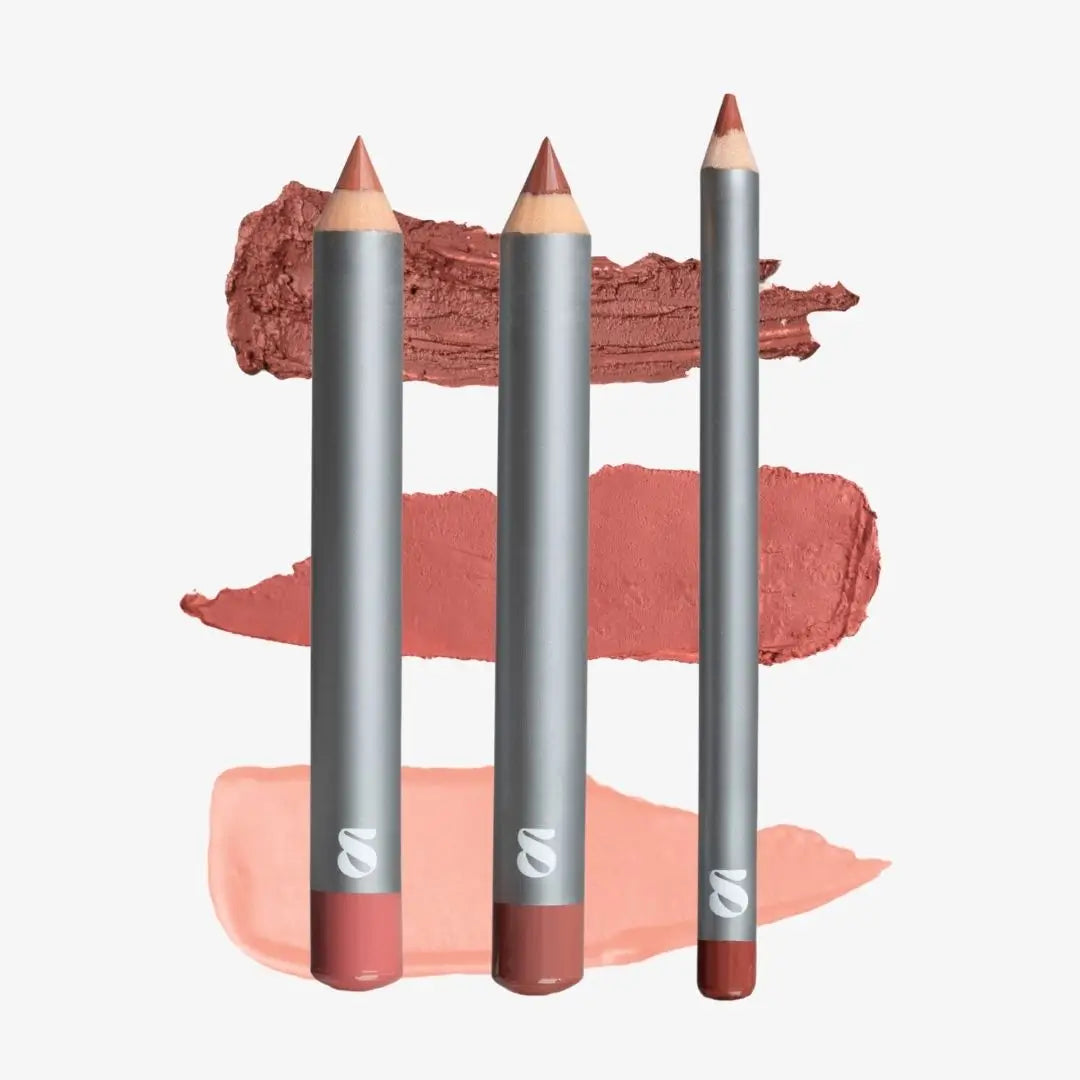 Three lip crayons with metallic silver bodies and pointed tips, arranged vertically in front of three horizontal makeup swatches in dusty rose, terracotta, and peach tones on a light gray background. Shade Creamy