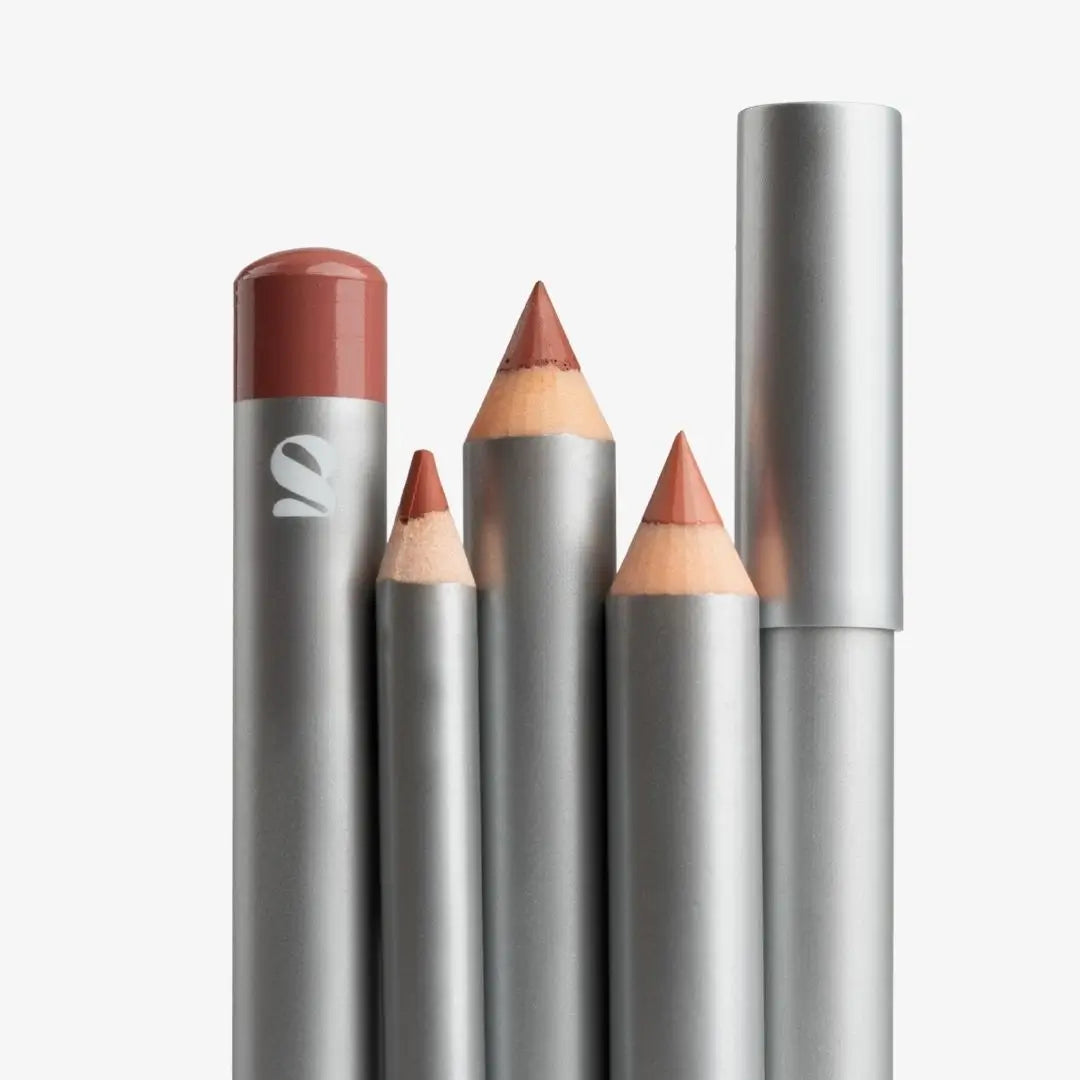 Four silver cosmetic pencils and a matching cap for a liquid lip product, arranged upright on a clean white background. One pencil is uncapped to show the tip, and the exposed formula is a muted rosy nude shade. Shade Creamy
