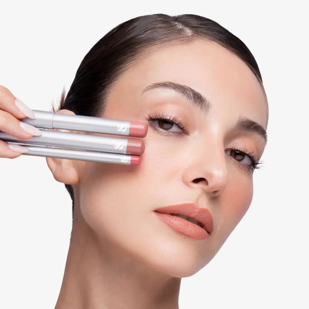 A close-up of a woman holding three nude pink lip liner pencils against her face, showing the product in use on a clean white background. Shade Creamy